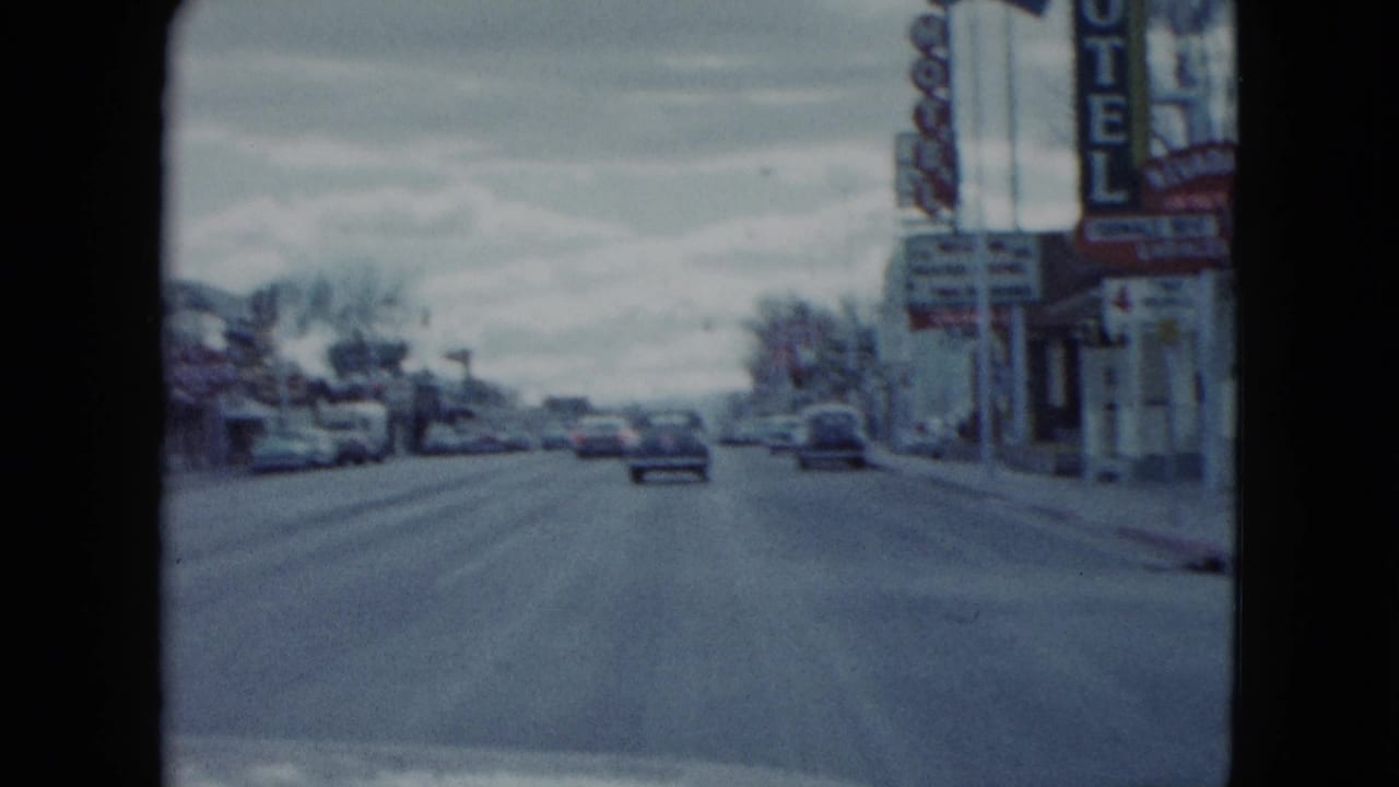1959 Nevada home-movie view down a small-town road with a car ahead, motel signs on the right, and roadside businesses.
