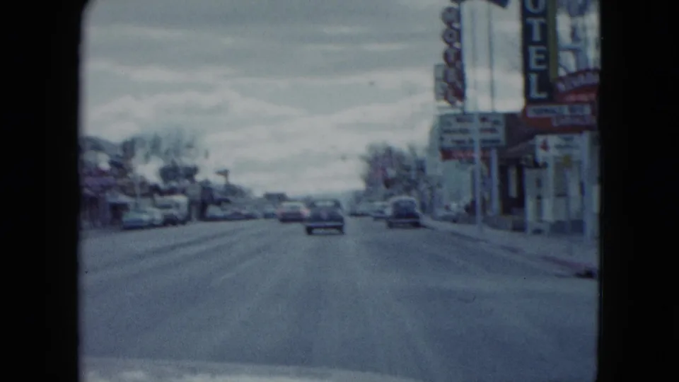 1959 Nevada home-movie view down a small-town road with a car ahead, motel signs on the right, and roadside businesses.