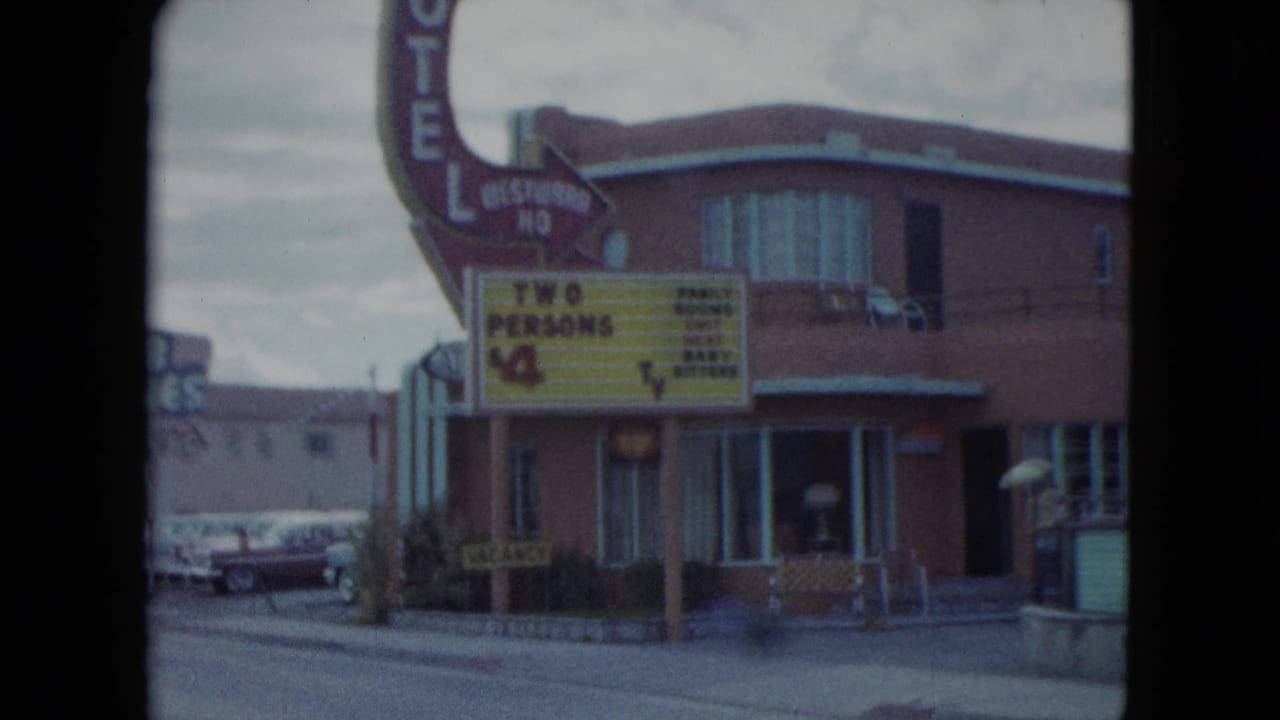 1959 Nevada 8mm view of a motel exterior with a prominent rate sign, sidewalk, storefront windows, and classic cars nearby.