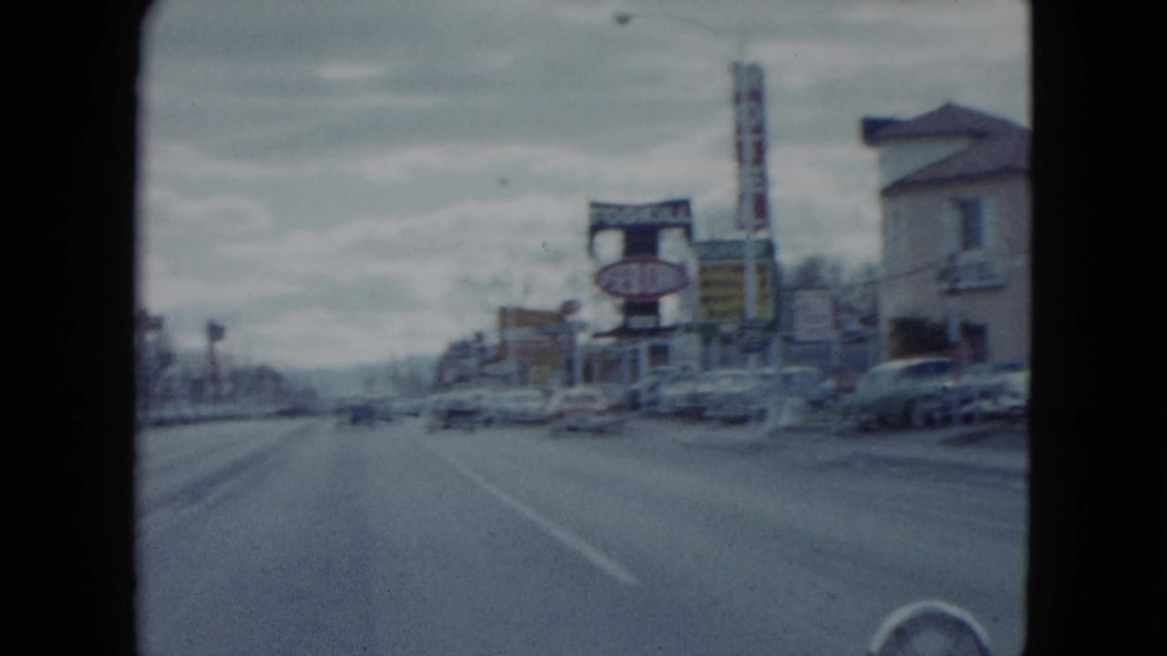 Car approaches a wide commercial street with motel and used-car signs under an overcast sky.