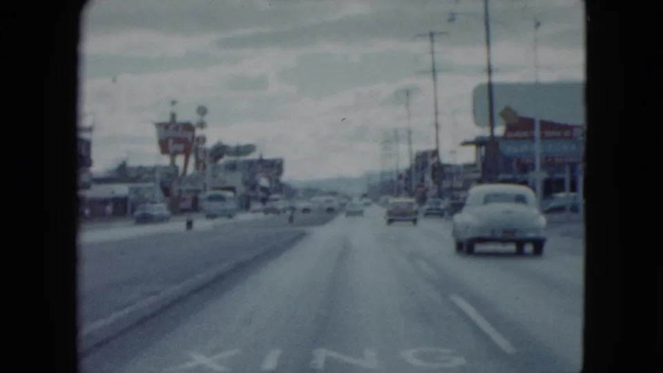 Moving 8mm street view in Nevada, 1959, showing classic cars, a raised roadside sign, utility poles, and a wide divided roadway.
