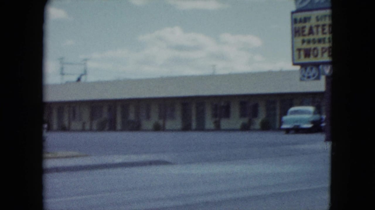 1959 Nevada roadside motel view with a long low building, large amenity sign, and a parked mid-century car near the curb.
