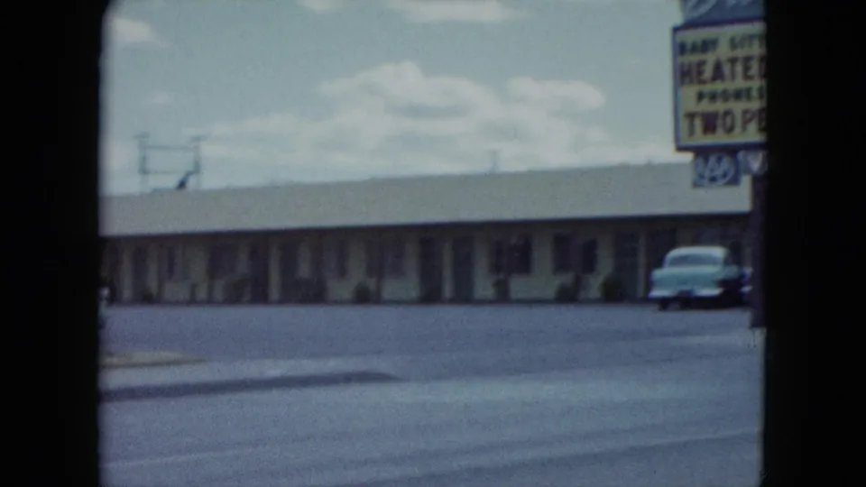 1959 Nevada roadside motel view with a long low building, large amenity sign, and a parked mid-century car near the curb.