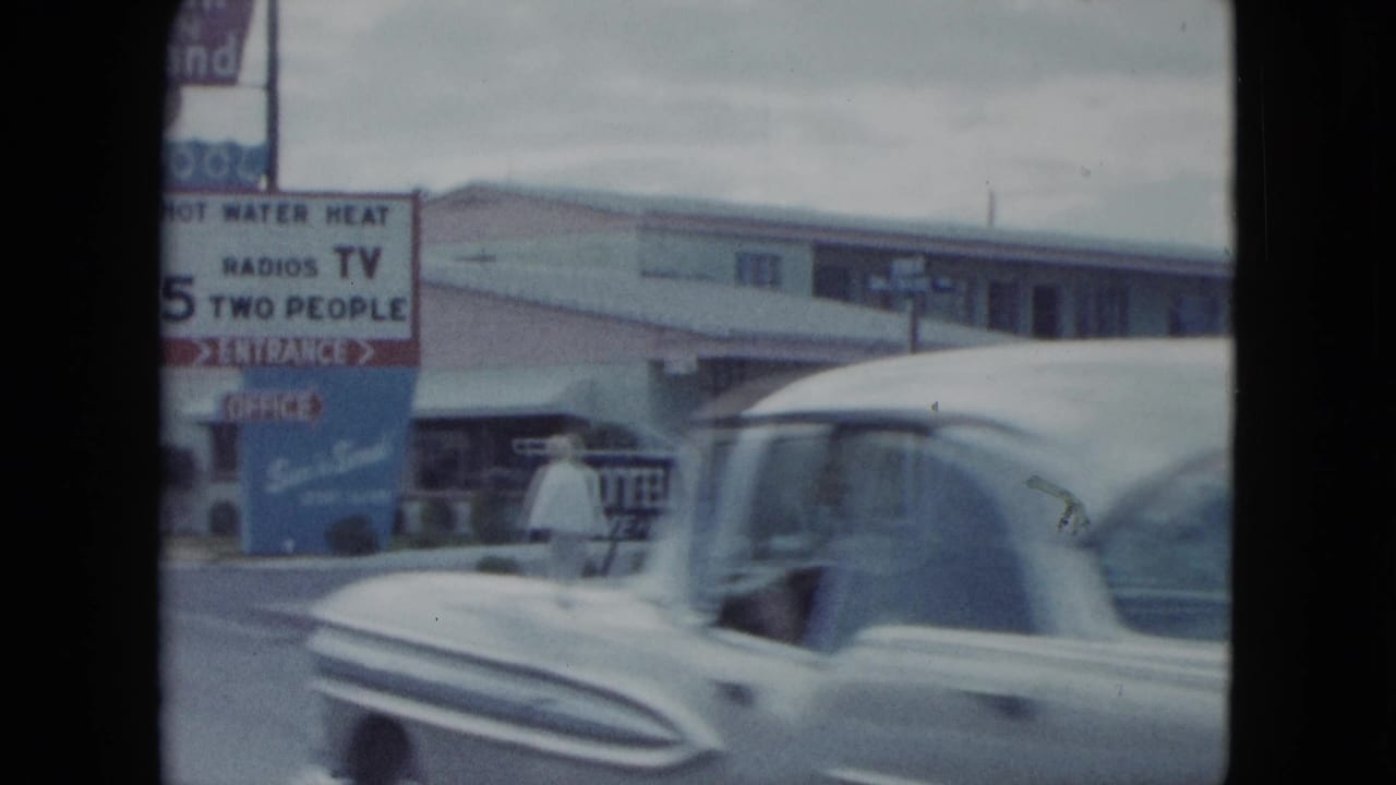 Passing mid-century car crosses the foreground of a 1959 Nevada motel view with a walking figure and roadside signs behind it.