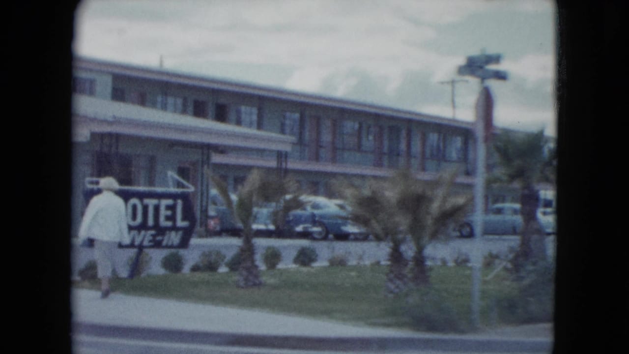 1959 Nevada motel frontage with a person walking by a drive-in sign, small palms, parked cars, and a two-story motel building.