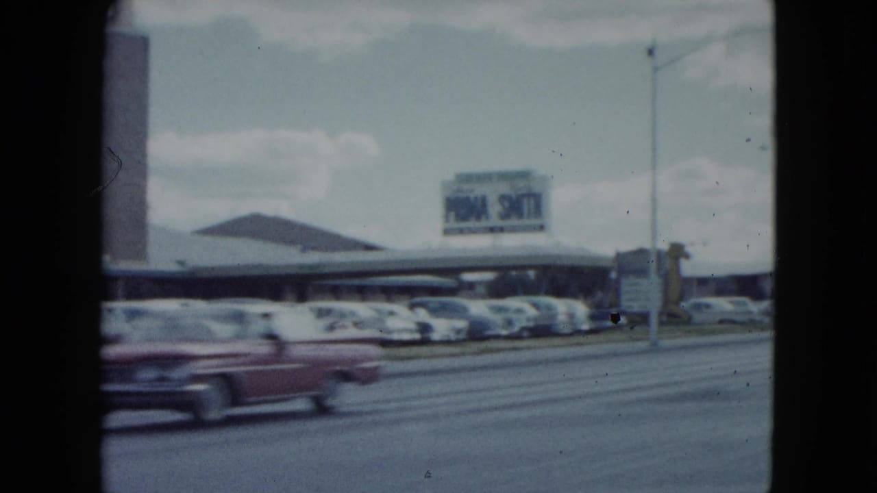 1959 Nevada frame showing the Sahara parking area, roadside signage, rows of cars, and a red classic car in motion.