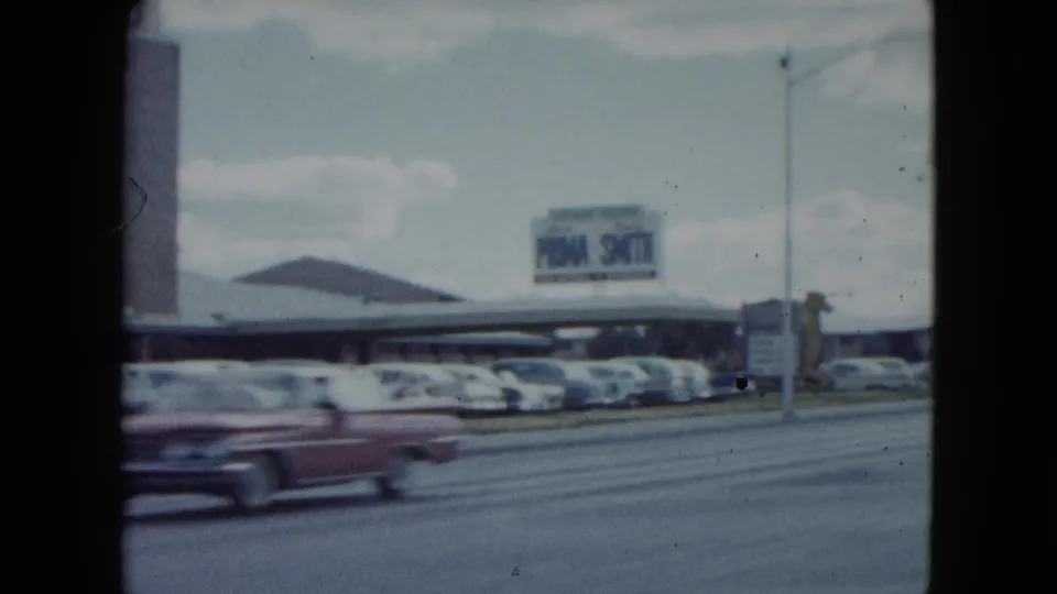 1959 Nevada frame showing the Sahara parking area, roadside signage, rows of cars, and a red classic car in motion.