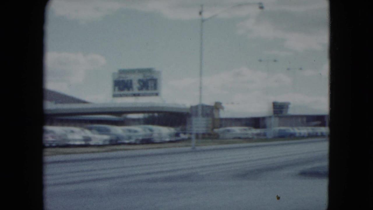 1959 Nevada road scene with late-1950s cars, commercial buildings, a distant sign, and soft 8mm film edges.