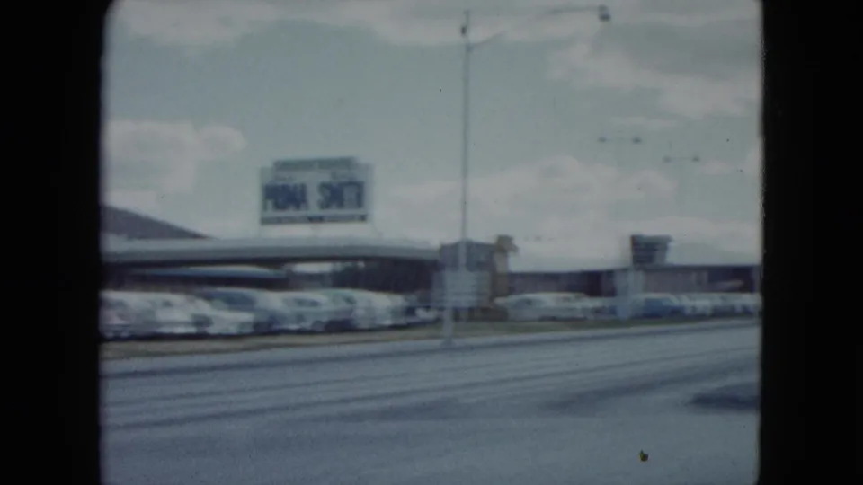 1959 Nevada road scene with late-1950s cars, commercial buildings, a distant sign, and soft 8mm film edges.