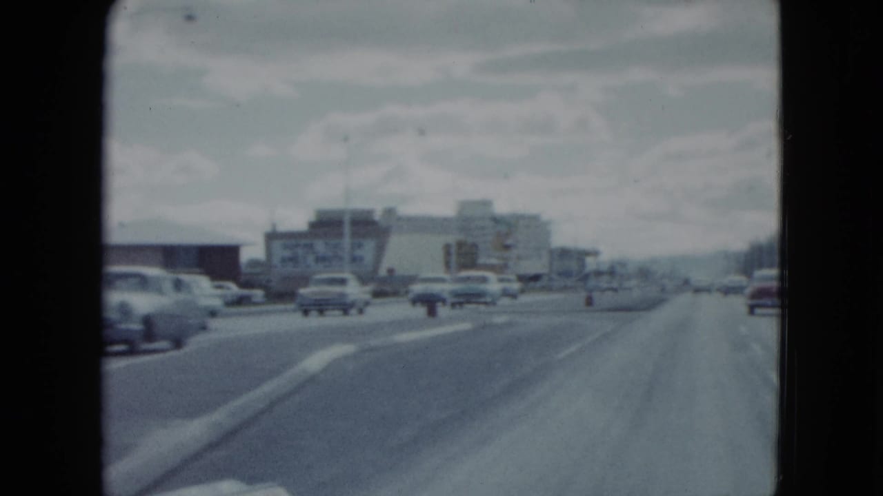 Forward-looking 1959 Nevada driving view with cars ahead, commercial buildings, roadside signs, and distant hills or skyline haze.