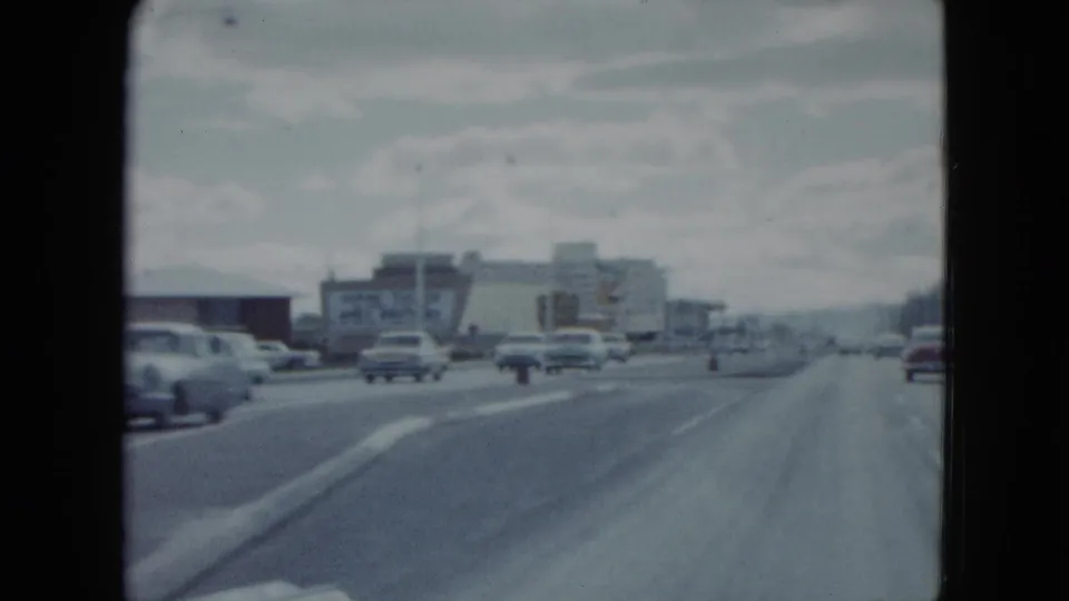 Forward-looking 1959 Nevada driving view with cars ahead, commercial buildings, roadside signs, and distant hills or skyline haze.