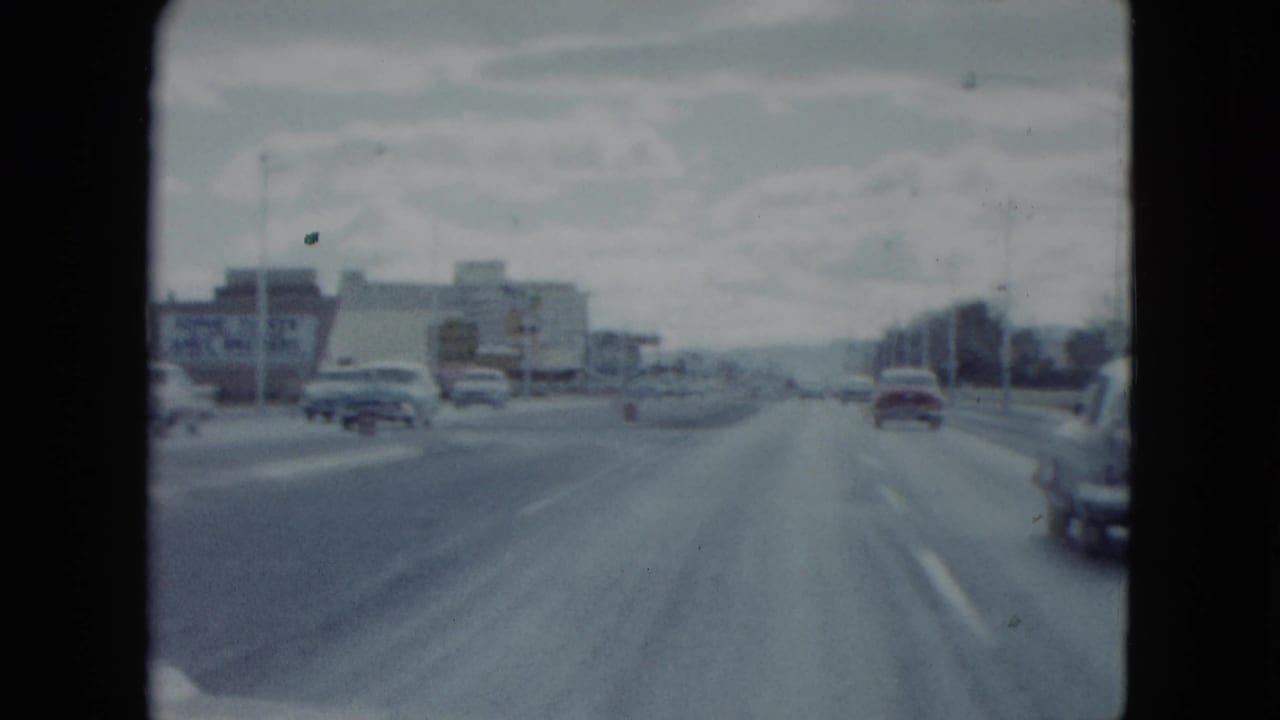 1959 Nevada 8mm view from a moving vehicle on a broad urban roadway with period cars, lane markings, and low roadside buildings.