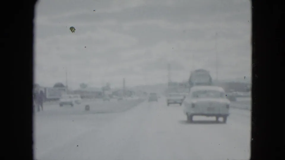 1959 Nevada road view with a rounded sedan ahead, lighter exposure, utility poles, and indistinct roadside traffic in the distance.