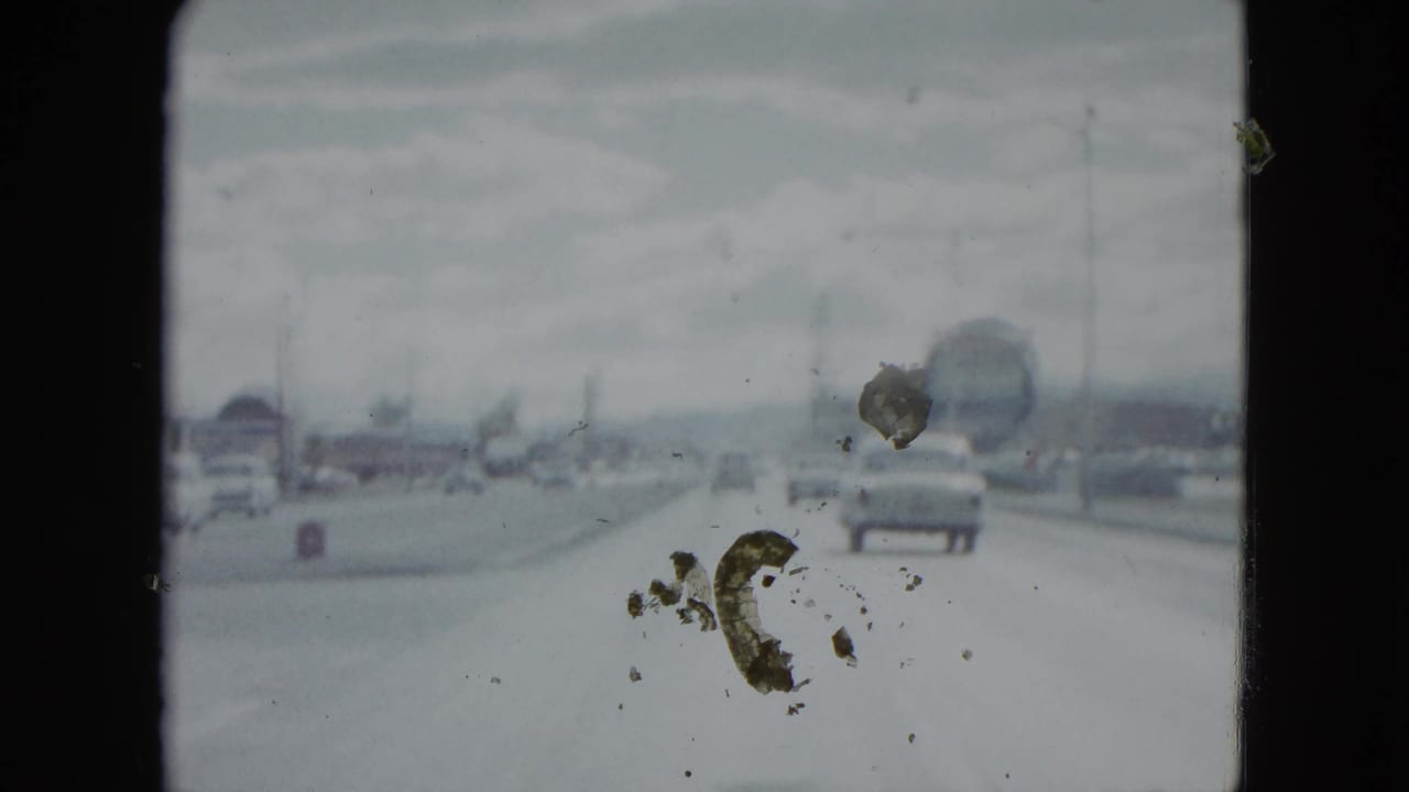 1959 Nevada 8mm frame showing a car ahead on a broad road, distant traffic, roadside structures, and prominent dust on the film image.