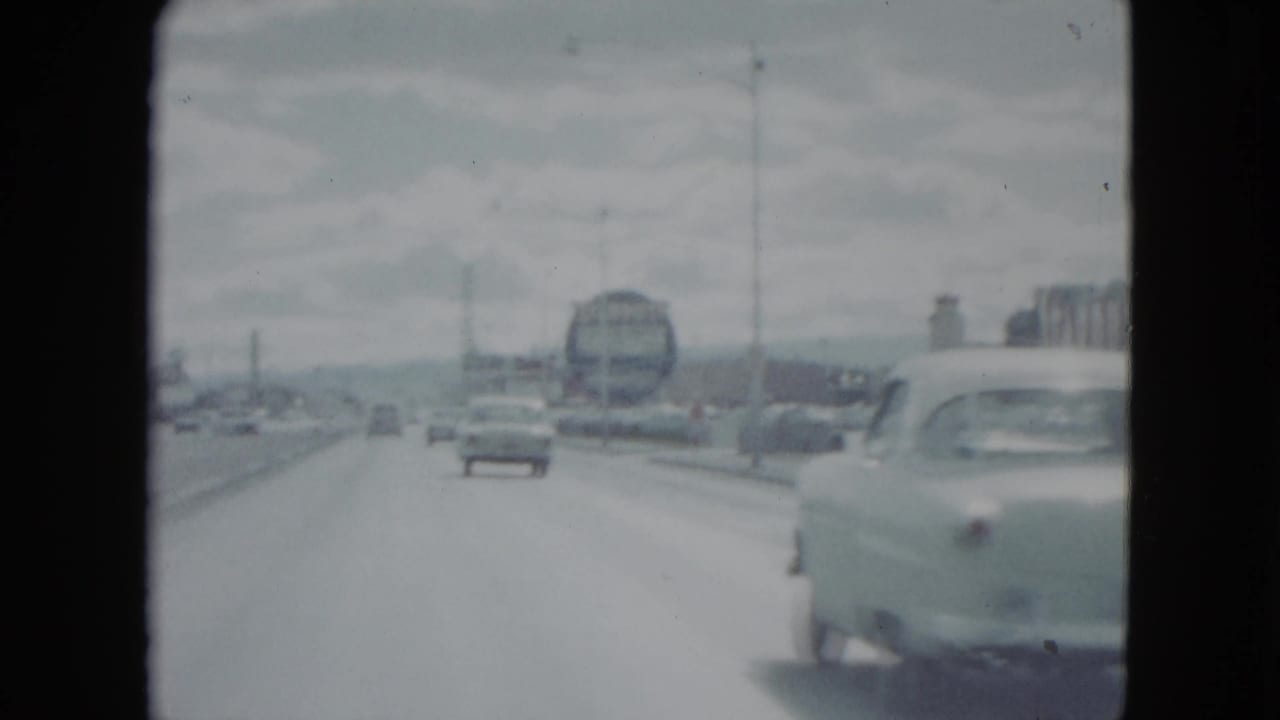 1959 Nevada roadway view with a bus-like vehicle ahead, light traffic, lane markings, utility poles, and a nearby car at the right edge.