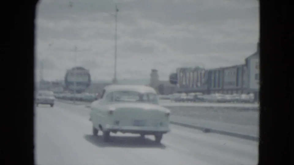 1959 Nevada 8mm frame showing a car on a wide road near commercial buildings and large roadside signage.