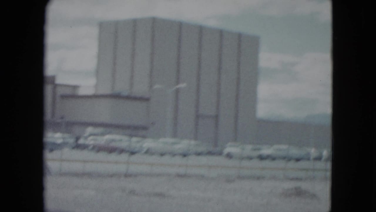 1959 Nevada home-movie frame showing a large commercial building, parked mid-century cars, fence, and cloudy sky.