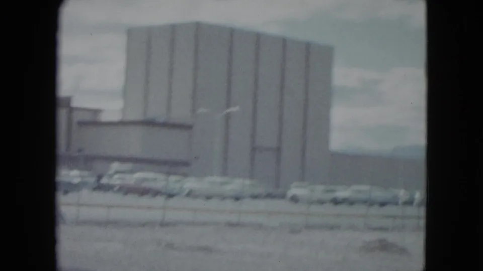 1959 Nevada home-movie frame showing a large commercial building, parked mid-century cars, fence, and cloudy sky.