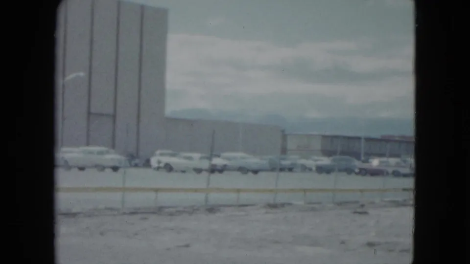 1959 Nevada frame with a tall building face, parked automobiles, low fence, and distant commercial structures.
