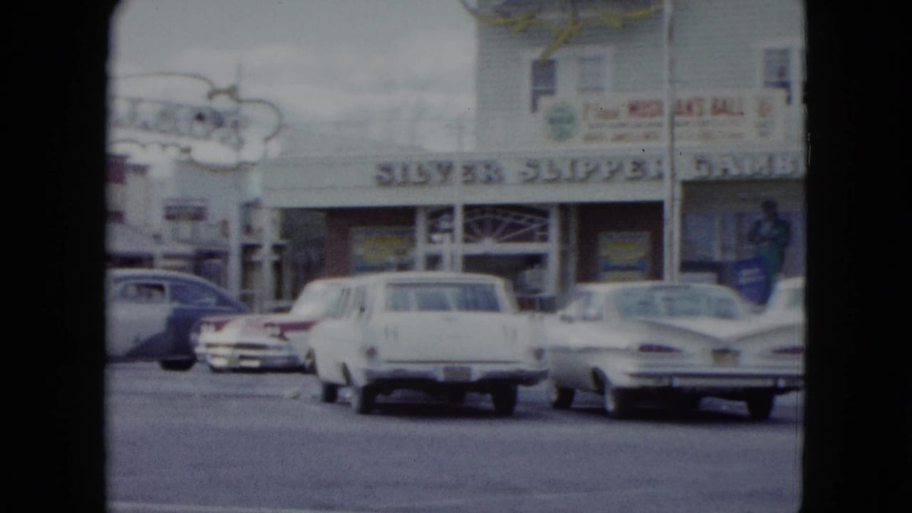 1959 Nevada home-movie frame showing parked cars along a street in front of local storefronts with readable signage.