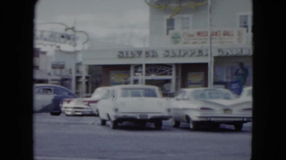 1959 Nevada home-movie frame showing parked cars along a street in front of local storefronts with readable signage.