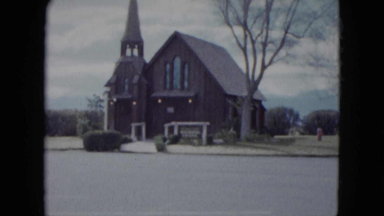 1959 Nevada home-movie frame showing a small dark wooden church with a steeple, trees, shrubs, and a quiet street in front.
