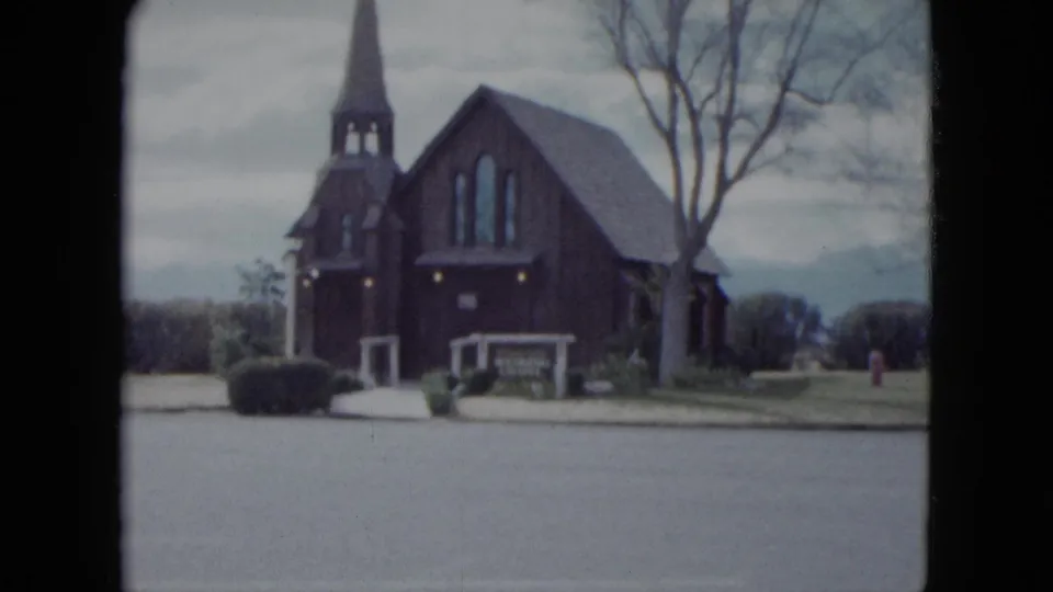 1959 Nevada home-movie frame showing a small dark wooden church with a steeple, trees, shrubs, and a quiet street in front.