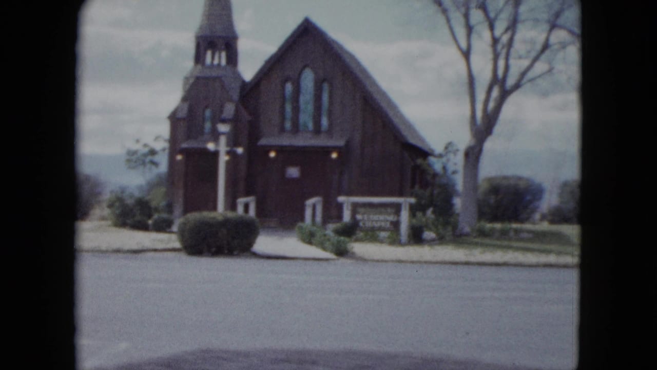 1959 Nevada home-movie frame showing a wooden church or chapel with steeple, front sign, trimmed bushes, bare tree, and quiet street.