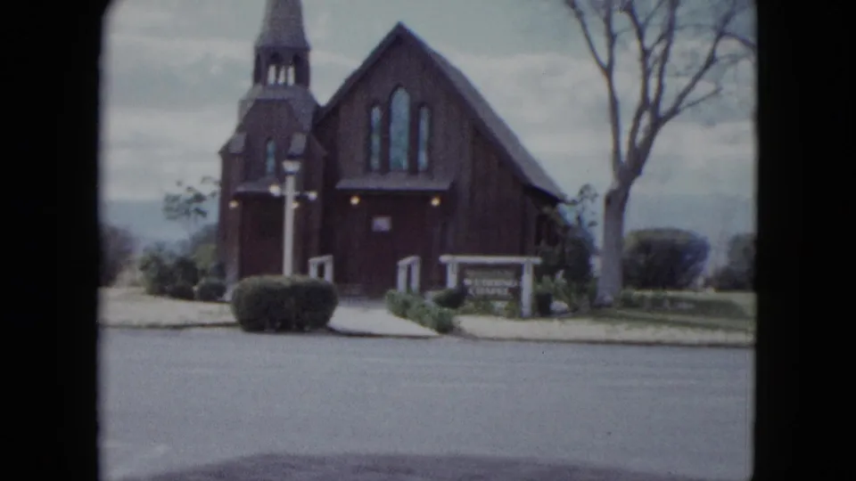 1959 Nevada home-movie frame showing a wooden church or chapel with steeple, front sign, trimmed bushes, bare tree, and quiet street.
