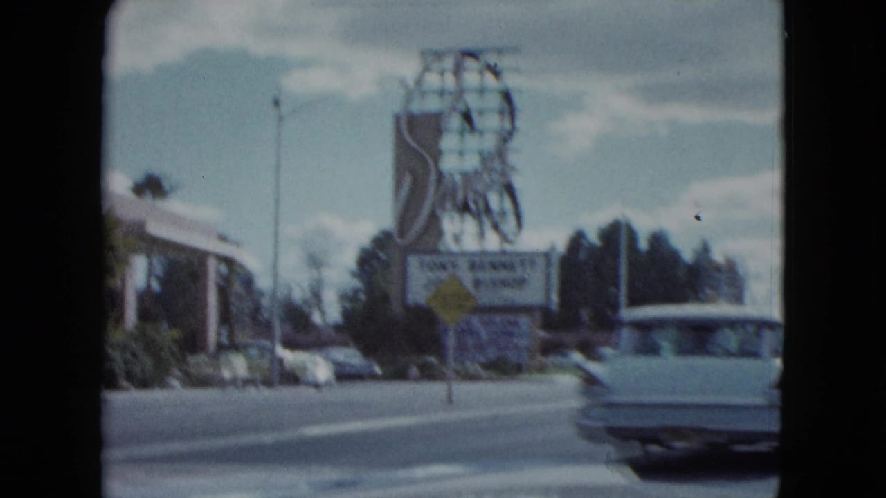 Large Sands roadside sign beside traffic, parked cars, and a passing classic car.