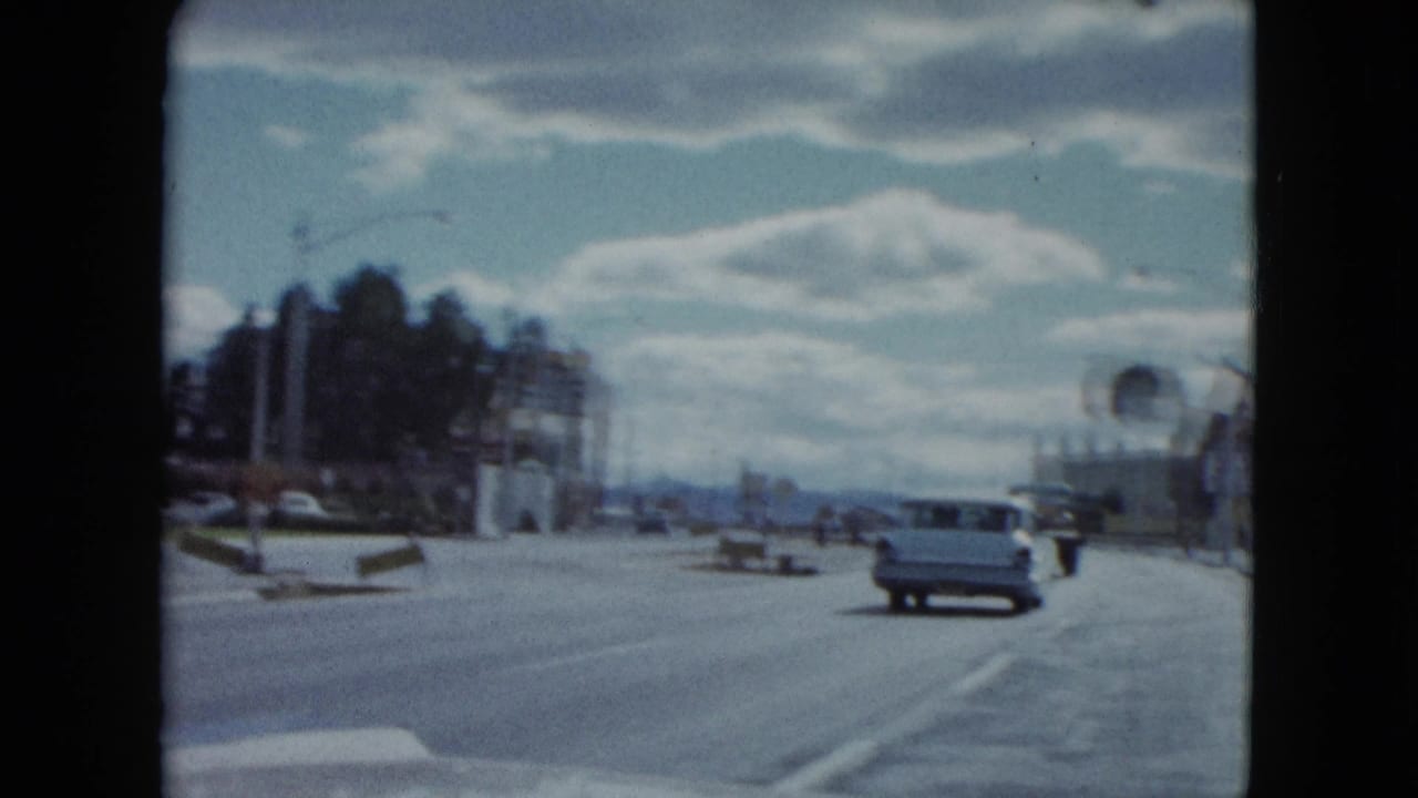 1959 Nevada home-movie view down a wide roadway with a classic car, roadside signs, trees, low buildings, and cloudy sky.