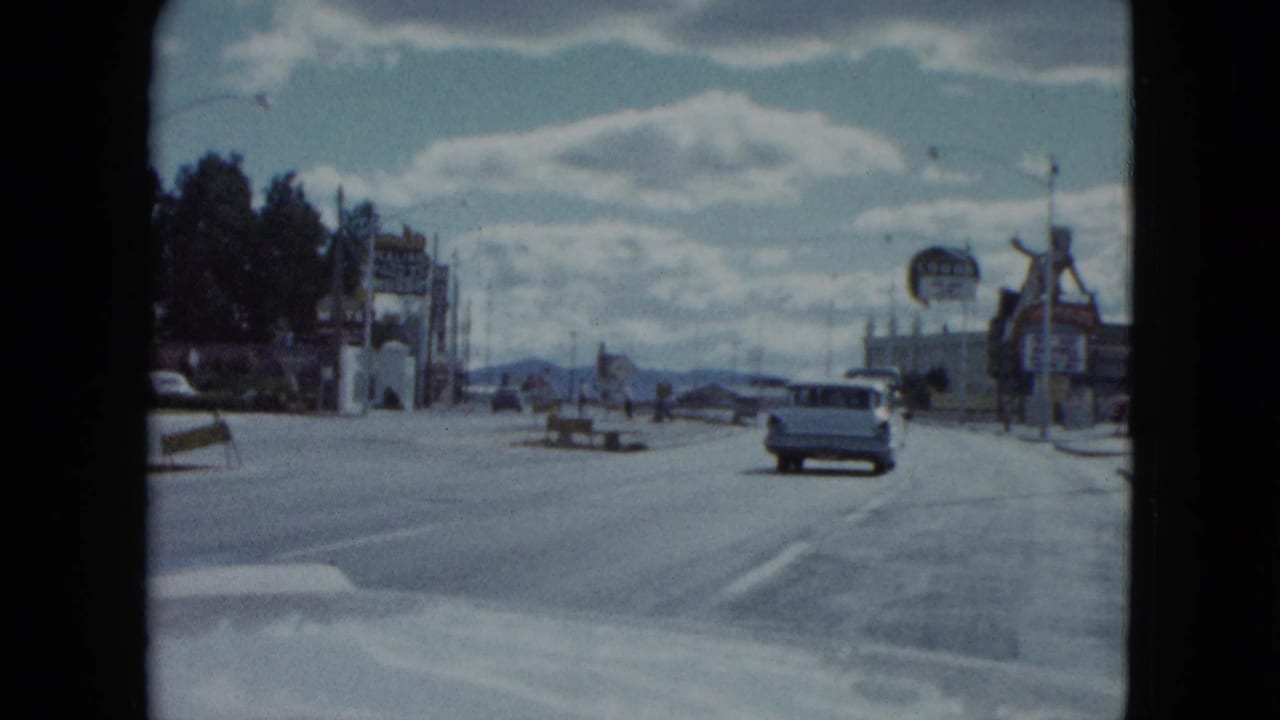 Wide street seen from a car with classic vehicles, roadside signs, and distant hills.