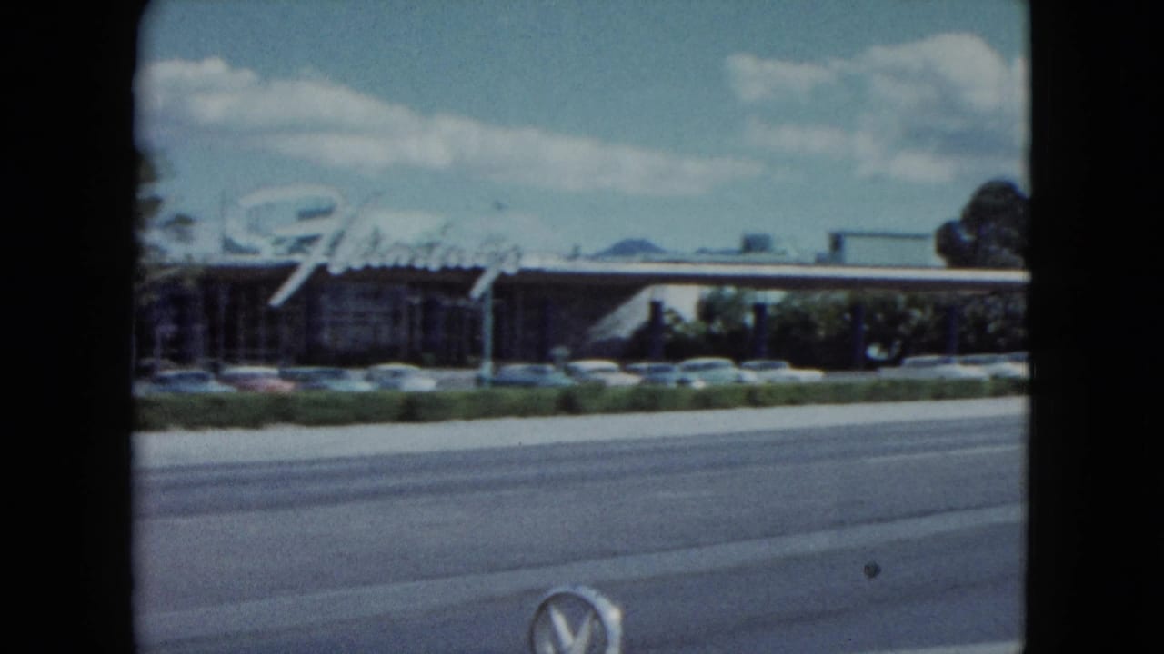 Roadside commercial building with a large script sign, parked cars, and a passing street.