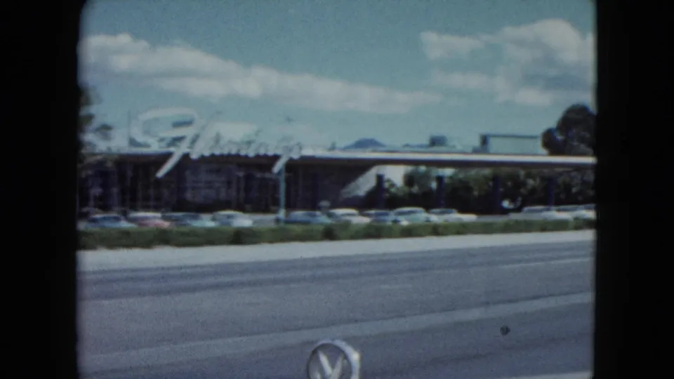 1959 Nevada roadside view of a low commercial frontage with a large script sign, parked cars, and a multi-lane street in front.