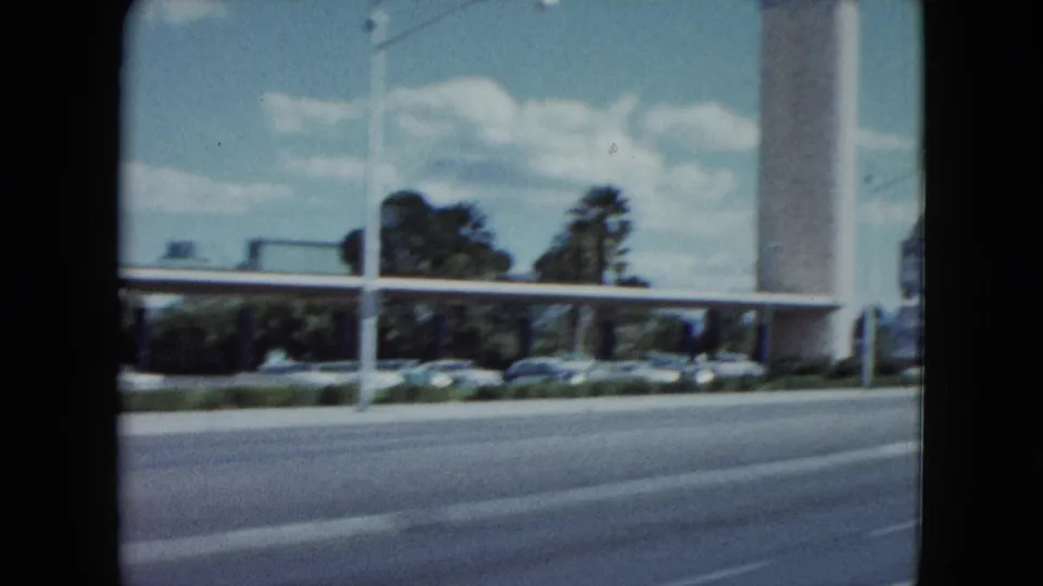 1959 Nevada street footage shows a road edge, parked cars, a tall white pole, low buildings, and trees under a bright sky.