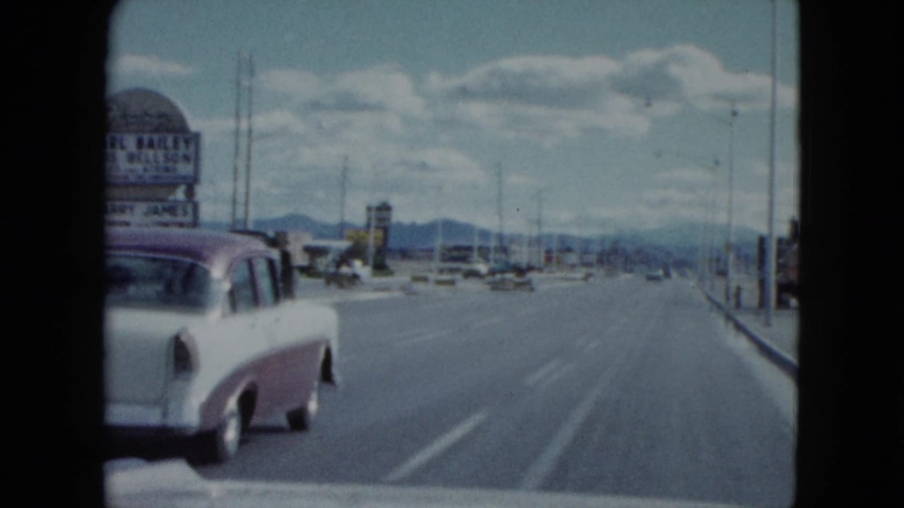 1959 Nevada home-movie view from a road with a white and maroon car, tall roadside signage, utility poles, and mountains in the distance.