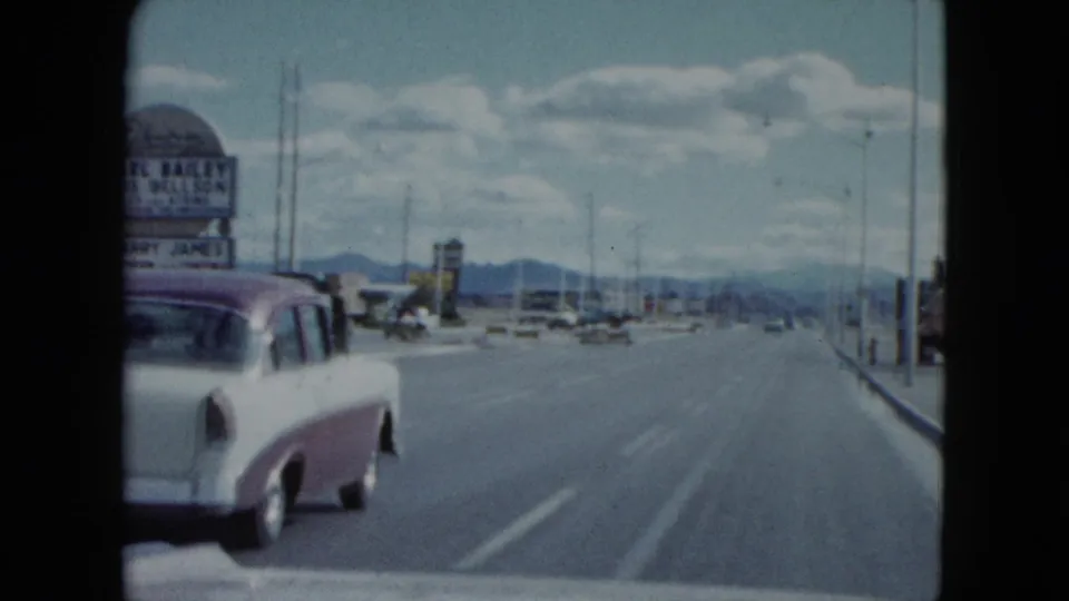 1959 Nevada home-movie view from a road with a white and maroon car, tall roadside signage, utility poles, and mountains in the distance.