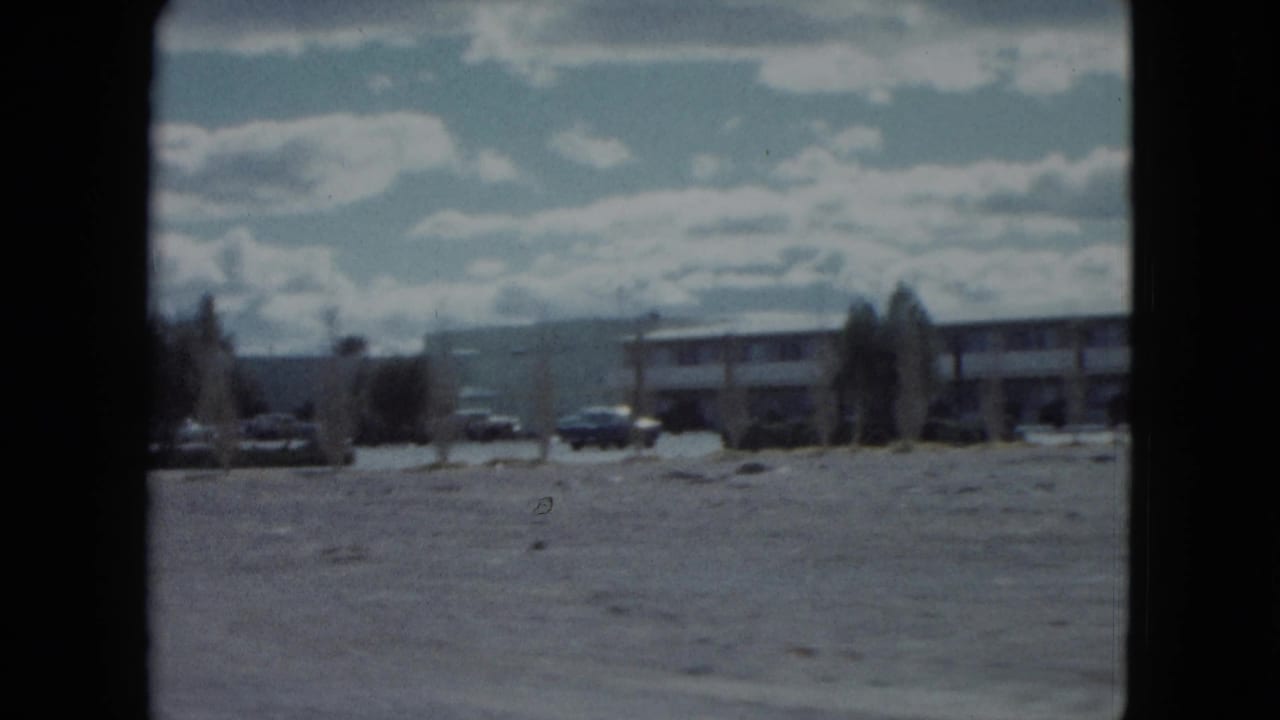 Low roadside building and parked cars seen across an open lot under a cloudy sky.