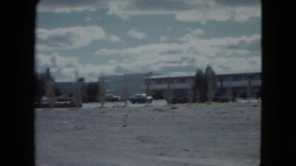 Brief 1959 Nevada roadside view of a low commercial building, parked cars, open ground, and mountains under bright clouds.