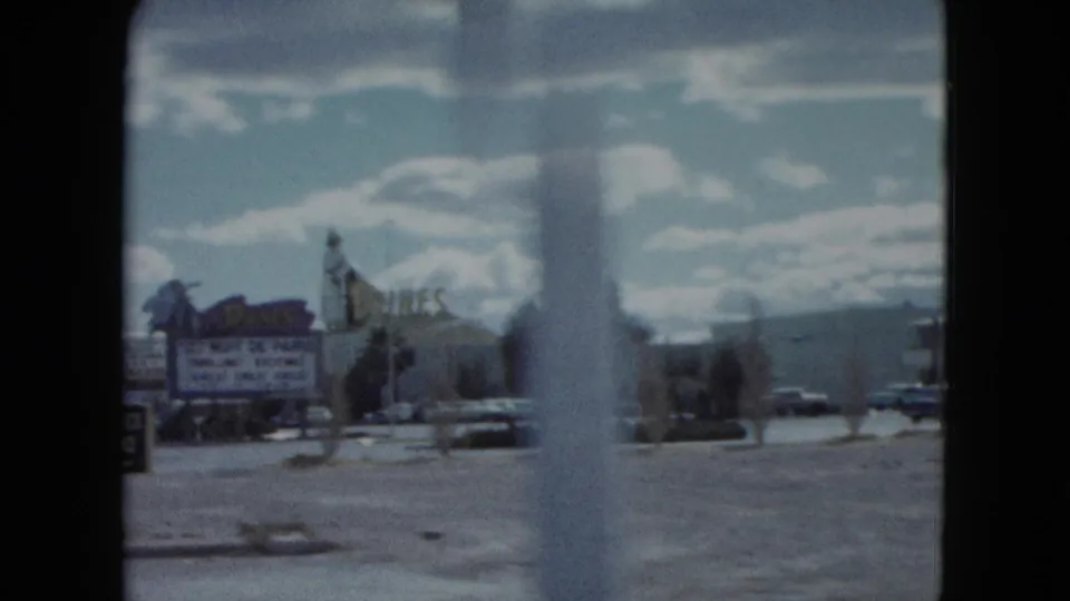 1959 Nevada home-movie frame with roadside signage, open parking, parked cars, and a blurred foreground pole or window edge.