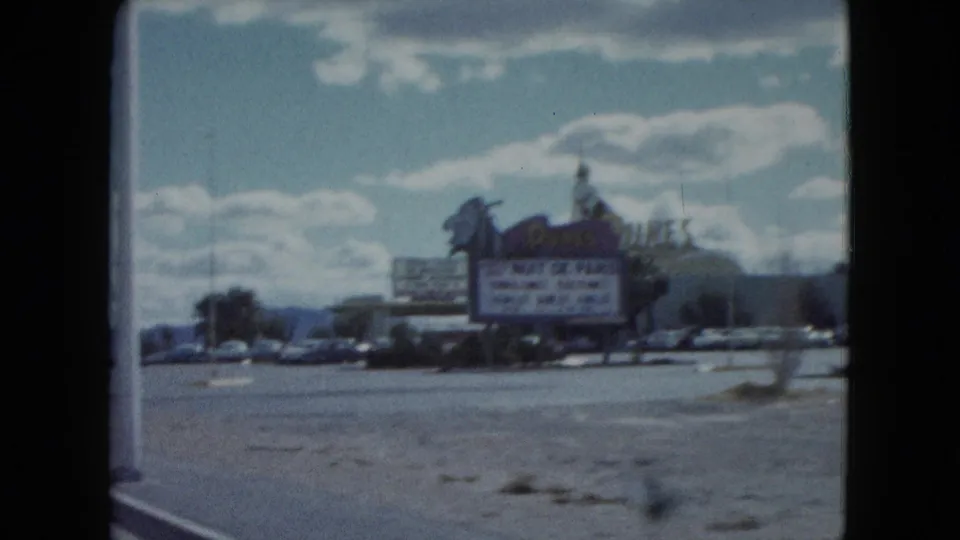 1959 Nevada road view passing the Dunes Hotel and Casino sign with a Nuit de Paris marquee, vintage cars, and distant mountains.