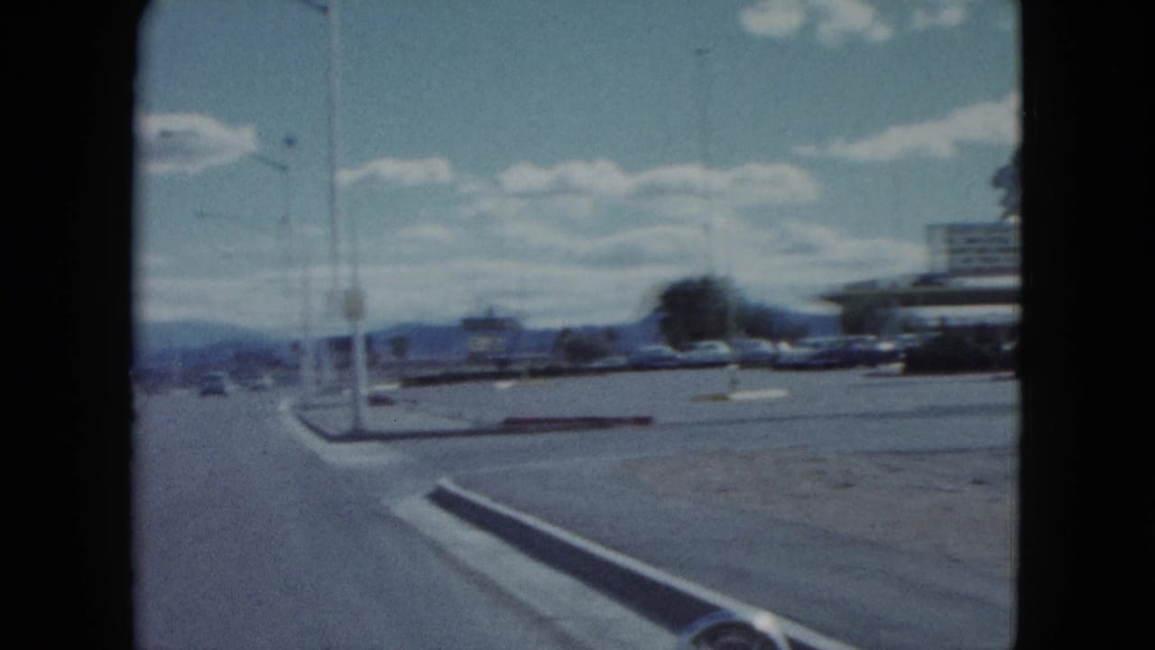 1959 Nevada road scene from a moving vehicle, with vintage cars, streetlights, curb islands, distant mountains, and roadside buildings.