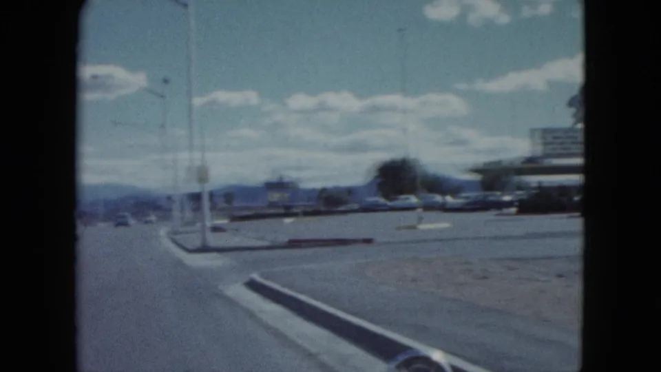 1959 Nevada road scene from a moving vehicle, with vintage cars, streetlights, curb islands, distant mountains, and roadside buildings.