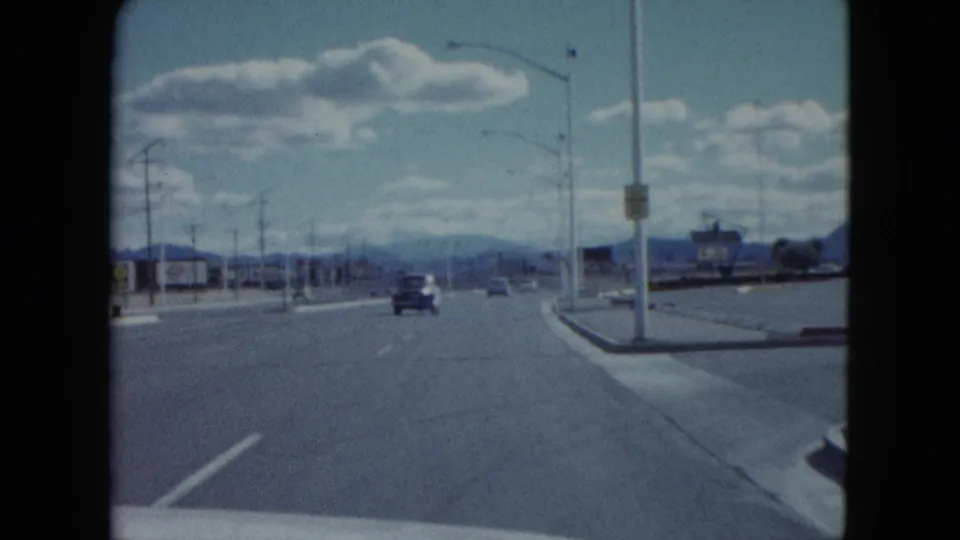 1959 Nevada driving view along a broad roadway with streetlights, roadside signs, a vintage car ahead, and mountains in the distance.