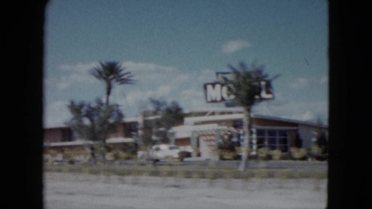 Motel building with large sign, palm trees, and a white car beside a desert roadside.