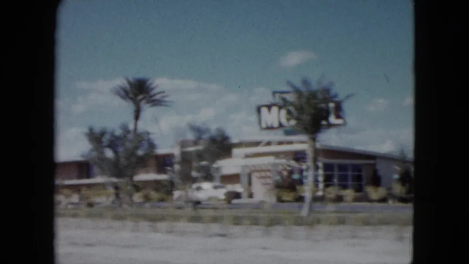 1959 Nevada home-movie frame showing a motel exterior, palm trees, and a white car near a roadside commercial strip.