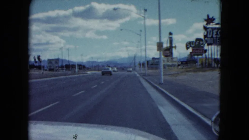 1959 Nevada windshield view down a broad highway with motel signs, cars, streetlights, and distant desert mountains.