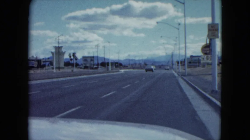 1959 Nevada road-trip frame looking along a multi-lane highway past commercial signage toward distant mountains.