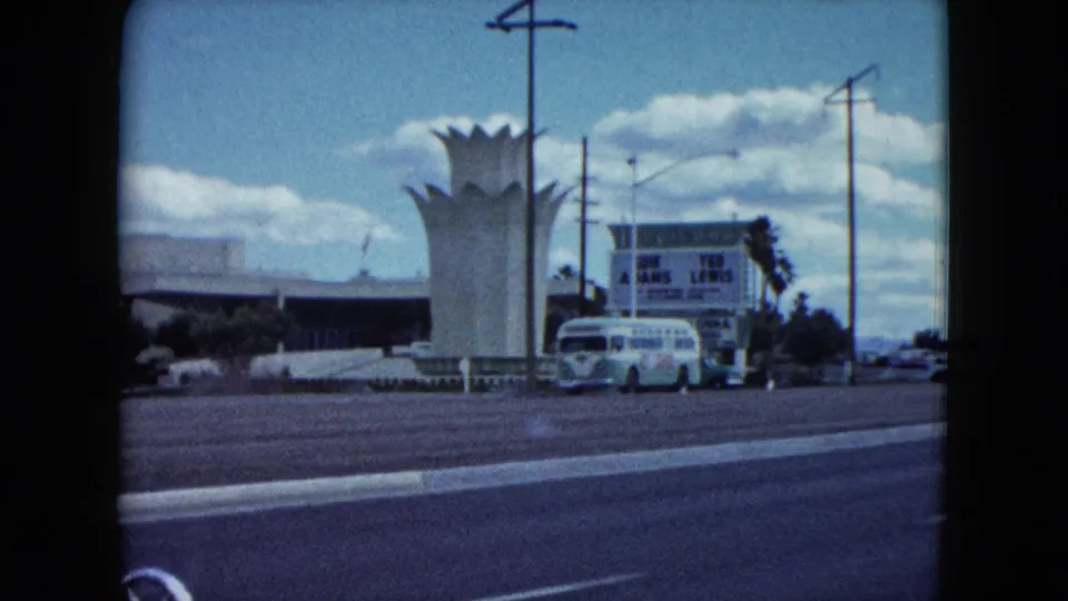 The drive-by view frames the white crown-like structure with a rounded bus, billboard lettering, poles, and road markings in 1959 Nevada footage.