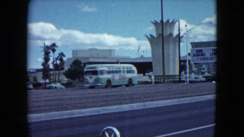 A 1959 Nevada roadside scene shows a pale bus passing the white crown-like structure, with a sign, low building, poles, and film gate edges visible.
